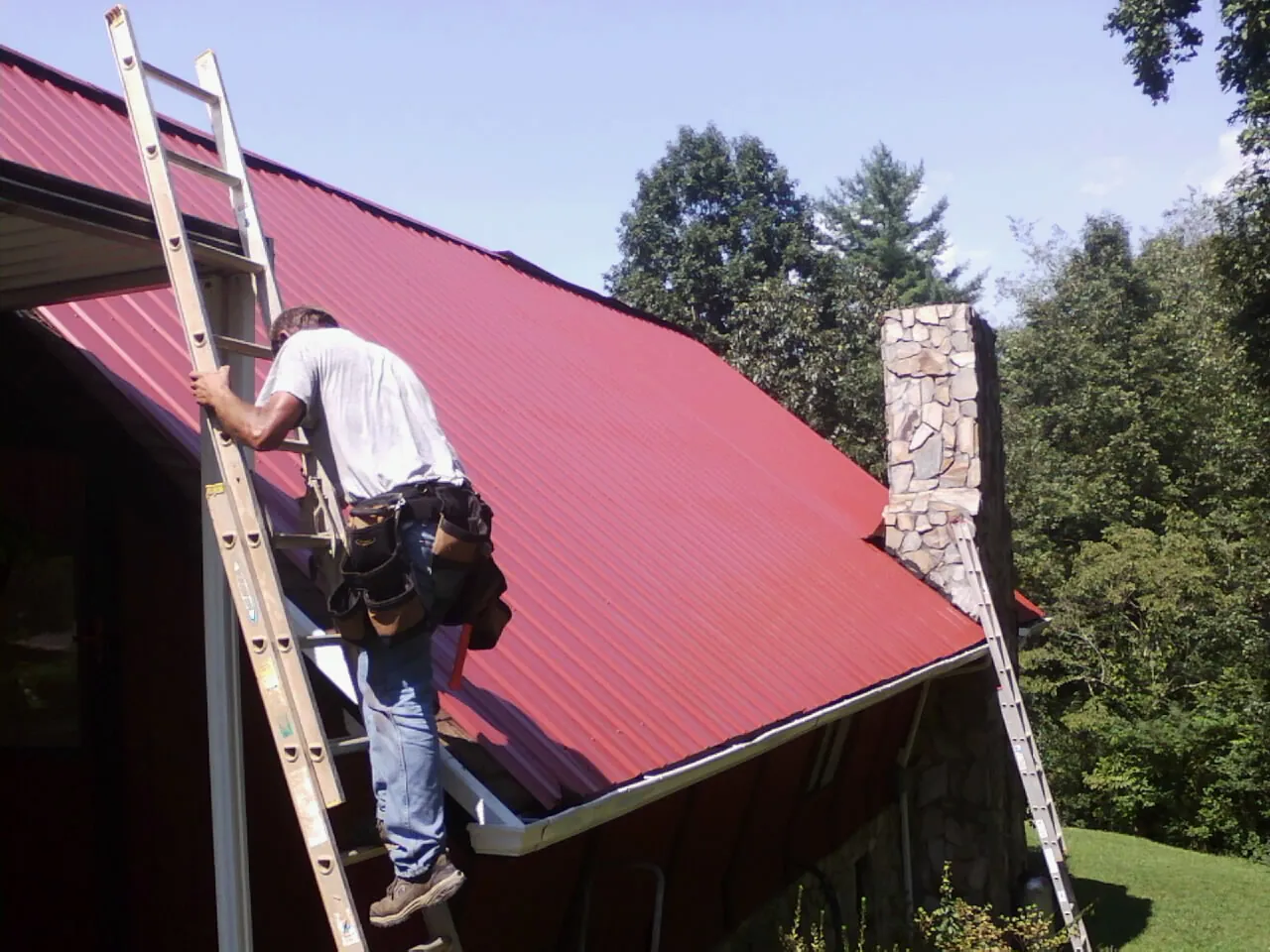 Roofer on a ladder beside a red metal roof and stone chimney — Soffit & Fascia Repair by Roofing Brothers in Branchport